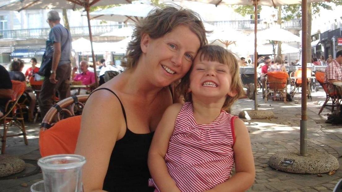 An old family photo of Mia and her mum Debby. Debby has short brunette hair and is wearing a black vest. Mia is a young child in the picture - she has short brunette hair and is wearing a red and white striped top. She is sat on Debby's lap. Mia has her eyes shut and a big smile on her face. Debby's head is against Mia's and she is looking at the camera and smiling. They are sat in an outdoor seating area of a bar/restaurant in a square, underneath a canopy of umbrellas.