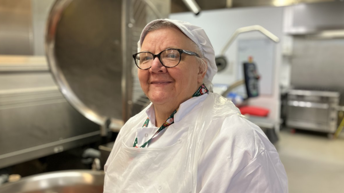 A woman wearing white overalls, a white chef's cap and a plastic apron, smiling to camera with large kitchen appliances in the background