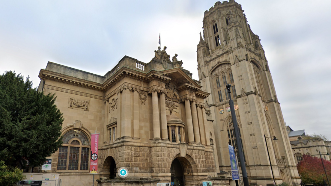 The front face of Bristol Museum and the Wills memorial Building in Bristol. There is a tree to the left of the museum and a flag outside advertising free entry to the museum.