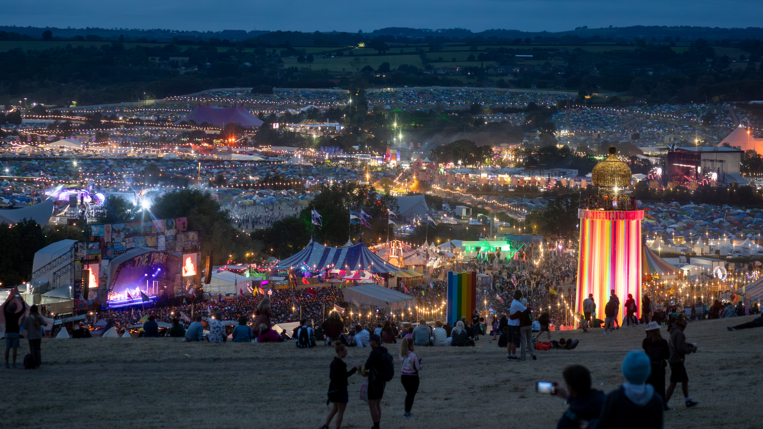 A view of Glastonbury Festival from the signs, with the Park Stage and Ribbon Tower in shot. There are lights illuminating the dark evening sky, with festival goers filling the grassy spaces.