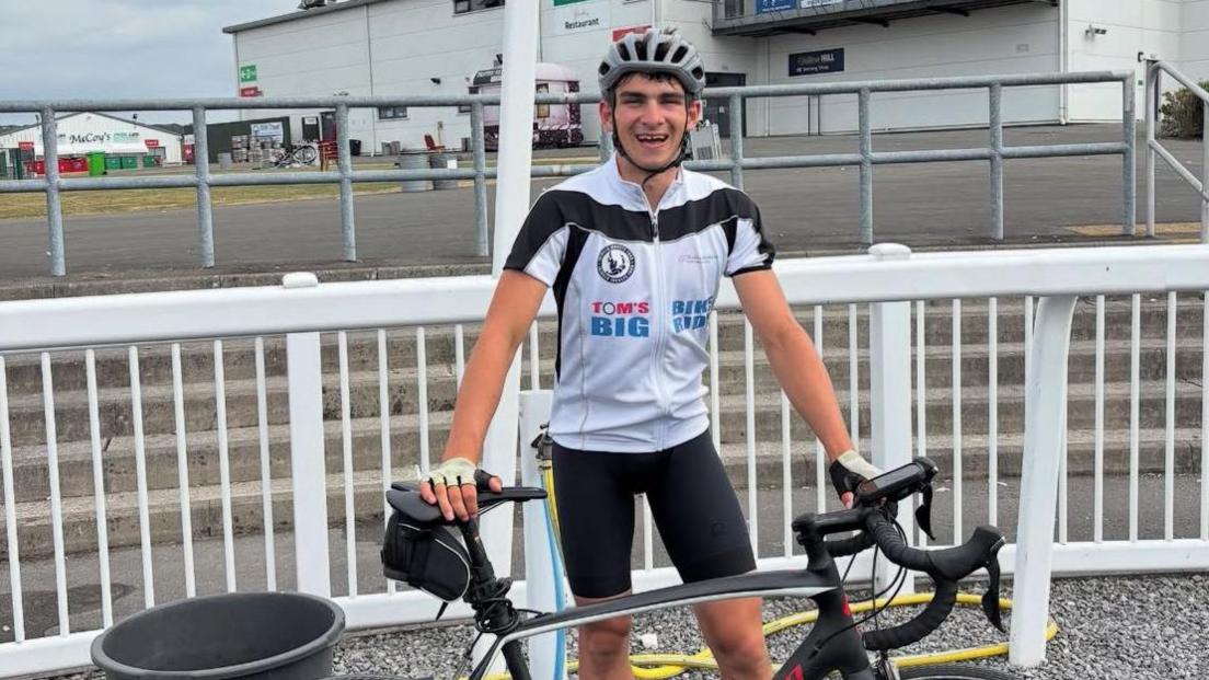 A smiling Tom Dunlop standing outside a racecourse wearing cycling gear and holding his bike.