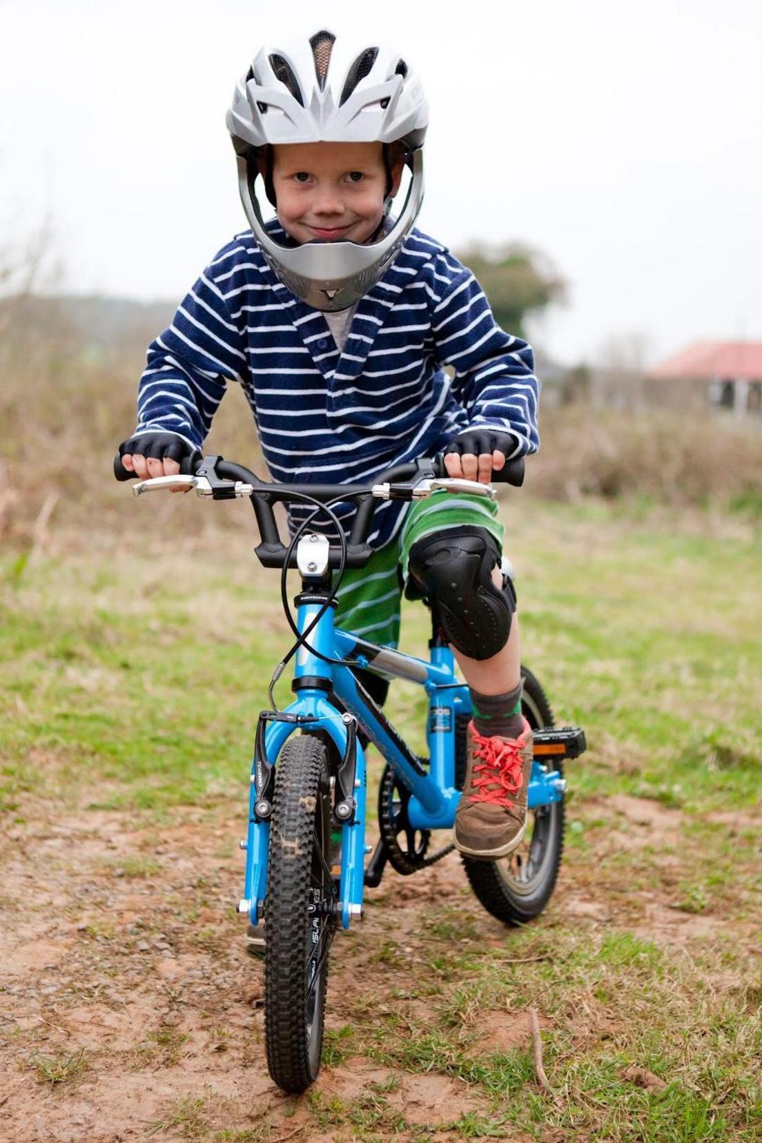 A young Ioan Hepburn in shorts and a striped long-sleeved top. He's stood on a blue bike, wearing knee pads and a full-face helmet.