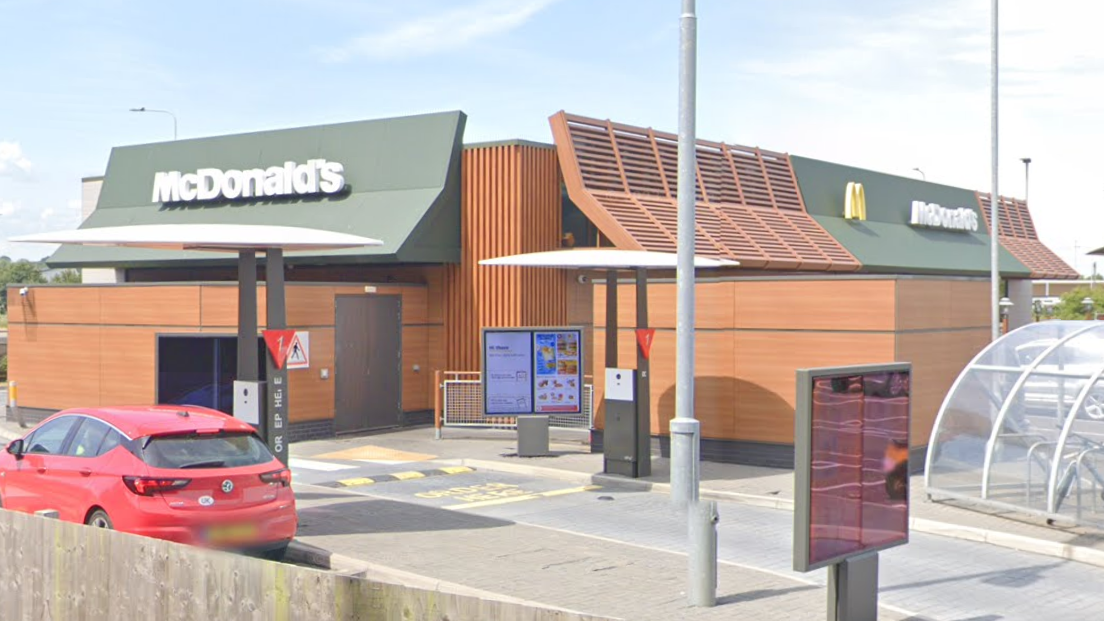 A McDonald's restaurant - a one storey building with brown exterior and a red car passing through one of its "drive through" lanes. A bicycle rack with a see-through canopy is on the right.