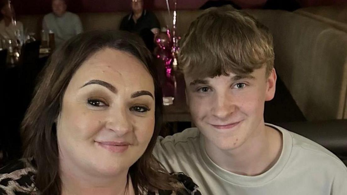 Clare Martin has shoulder length dark brown hair. She wears a leopard print top and smiles at the camera. Her son Alfie is next to her, smiling at the camera. He has light brown hair and is wearing a white top. 