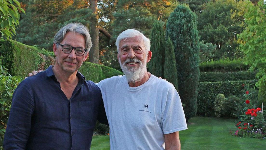 Two men standing in a garden with their arms around each other. One the left is a grey haired man wearing black-rimmed glasses and a blue shirt. On the right is a man with grey hair and a grey beard wearing a pale T-shirt marked on the left side of the chest with the letter "M".