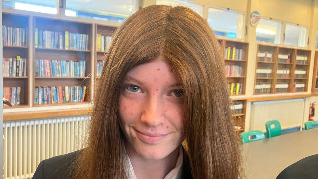 Kayleigh is wearing a black blazer and white shirt and has long red/brown hair parted in the middle