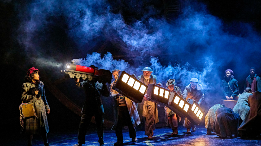 Actors carry an illuminated model train across a dark, smoky stage