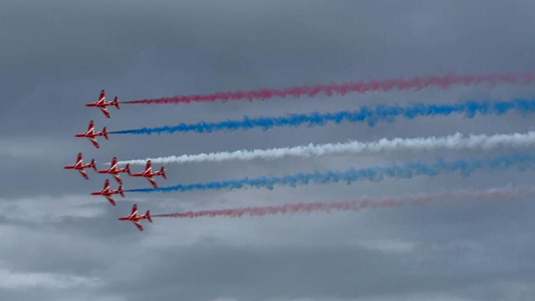 The Red Arrows in formation with red, white and blue smoke trailing behind the red planes.