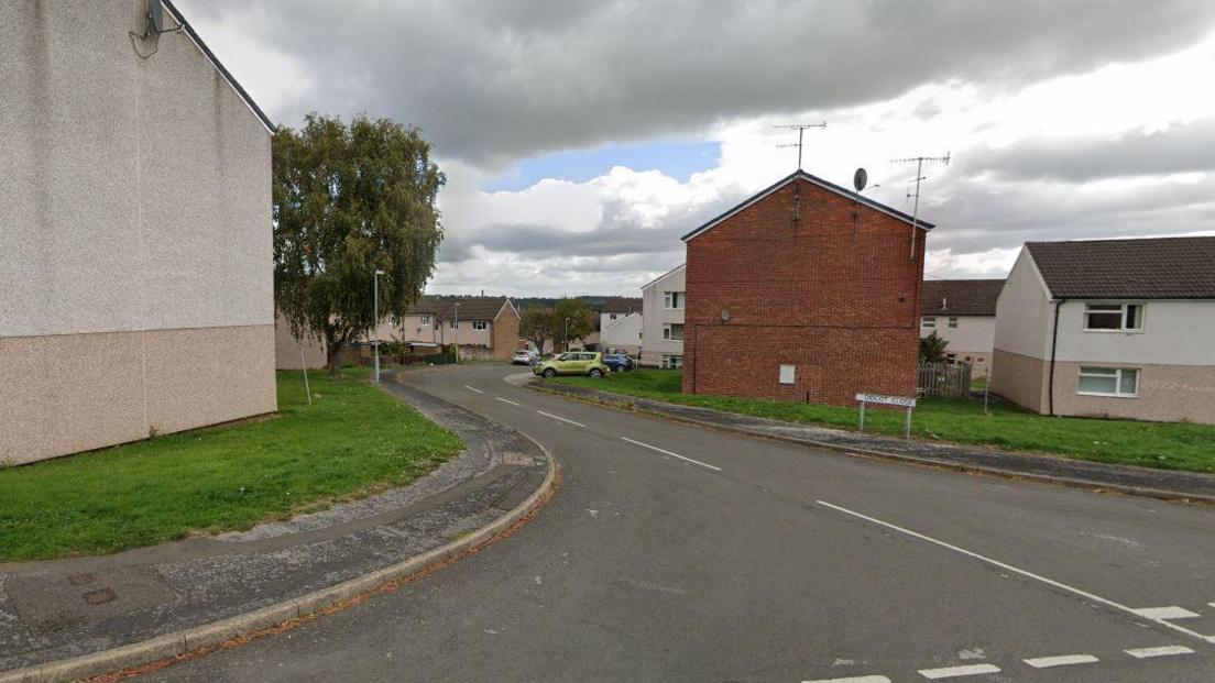 A Google street view of Didcot Close in Chesterfield. On the right, a street sign can be seen and in the distance are a number of buildings - possibly houses or flats - painted cream. 