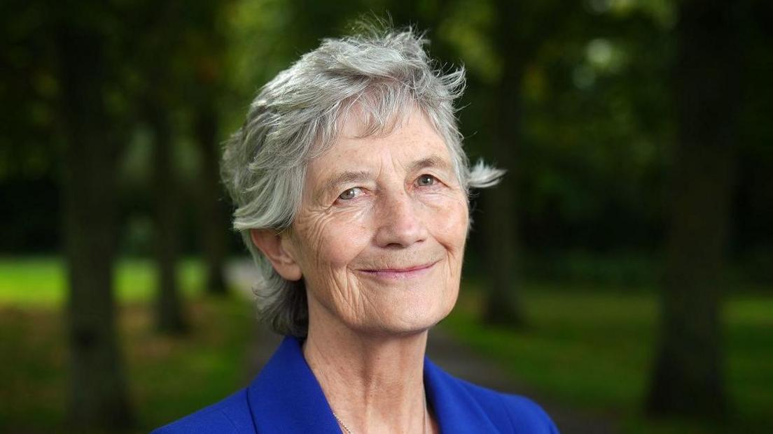Catherine Connolly has grey hair down to the bottom of her neck and is wearing a blue suit jacket. She is standing outside with a number of trees in the background.