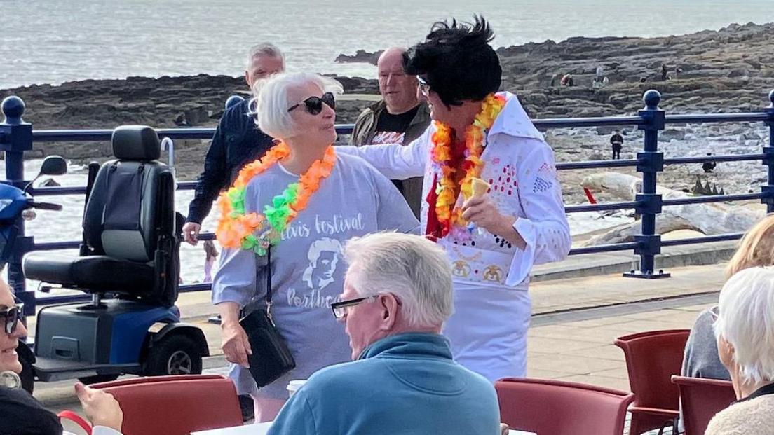 An Elvis with a flower necklace talks to a woman in an Elvis jumper