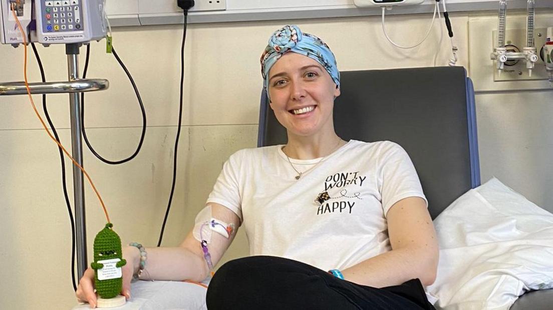 Vicky Green smiles as she sits in a chair receiving chemo treatment. She is holding a knitted green cactus toy. She is wearing a blue patterned head scarf.