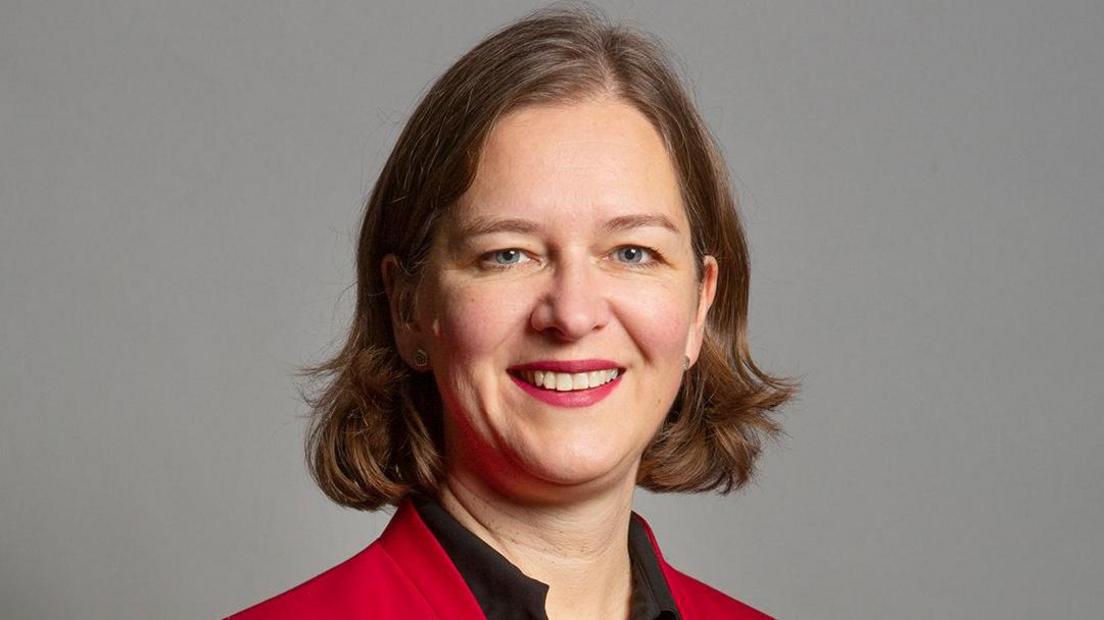 Fleur is standing against a professional looking grey wall, smiling.
The photo is cropped from shoulder height upwards.
She has blue eyes and light brown hair to her shoulders. 
She is wearing red lipstick and silver stud earrings.
She is wearing a black v neck blouse under a red collarless blazer. 