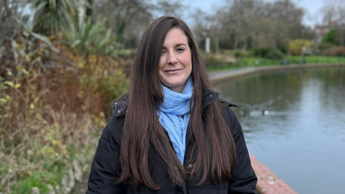 A woman with long brown hair, wearing a light blue scarf and black puffer jacket, stands on a path next to a lake.