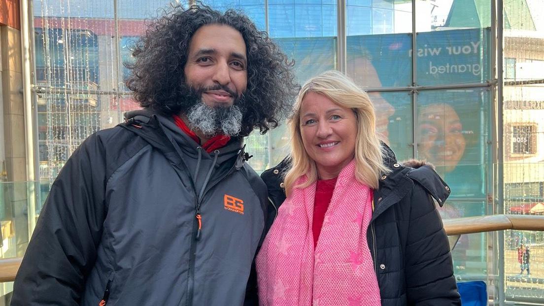 Kamran and Mandy are standing next to each other and smiling. Kamran is wearing a grey coat and has brown curly hair. Mandy has blonde hair and is wearing a red top and a pink scarf