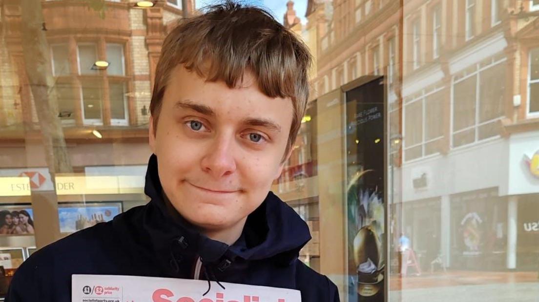  Head shot of Adam Gillman who has short blond hair and is standing in front of a shop window