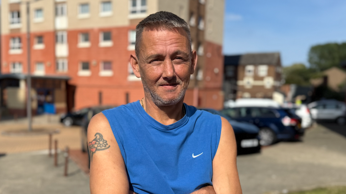 Geoff Bennett looks like he is in his 40s and is wearing a sleeveless blue vest top with a Nike tick on. He has a tattoo of a flower and heart on his right bicep. He has a grey hair and beard and has his arms crossed, and is squinting slightly into the sun.