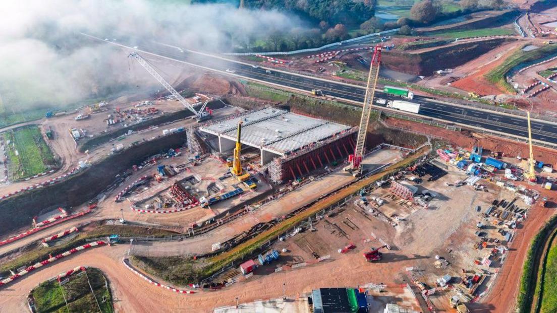 A bird's eye view of a building site next to a busy road. A large box-like structure is mid-construction a few metres from the carriageway. Several cranes are on the building site.