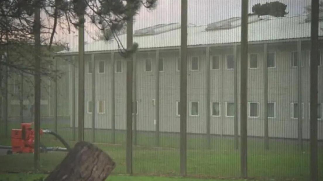 A grainy image of the former detention centre which has lots of windows and a slanted roof. There is a large fence with poles in front. 