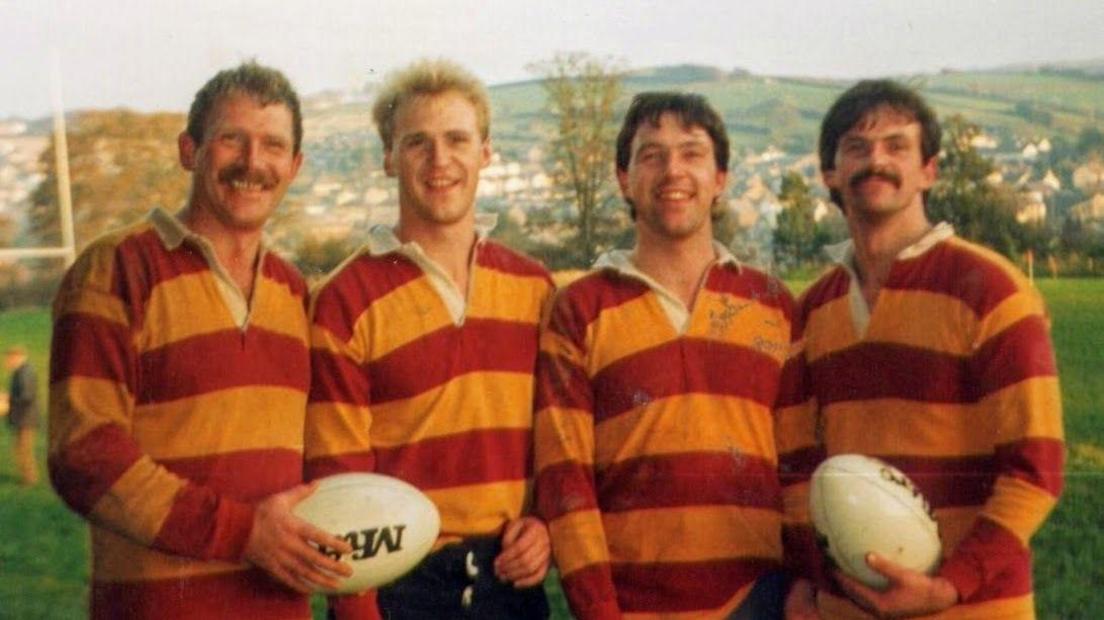 Rob Ewen stood on the left with his three brothers. They are wearing red and orange striped rugby tops and two of them are holding rugby balls. 