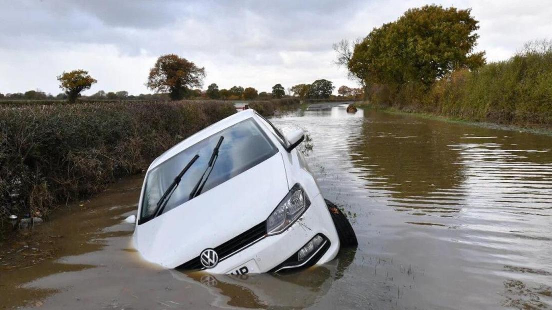 A white VW is partially submerged in floodwater - the water on the road resembles a river