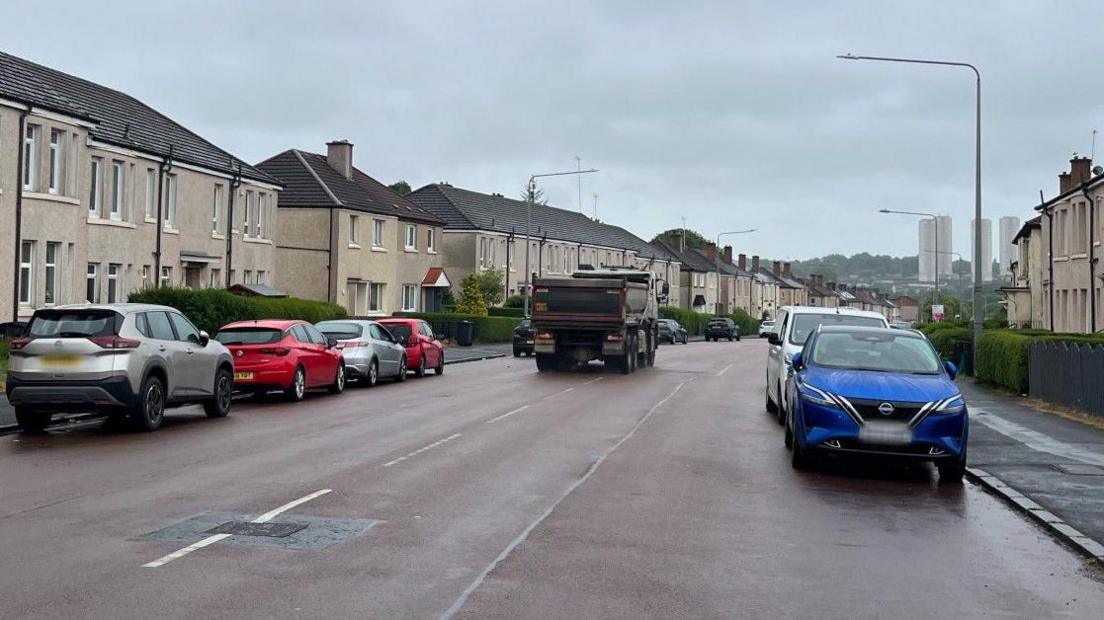 A street view of a wide main road with terraced homes on either side and vehicles parked along the pavement on both sides