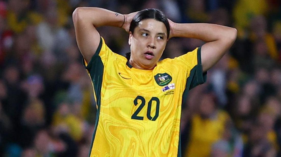 Sam Kerr wearing her Australia football shirt. It is predominantly yellow with dark green sides and the number 20 in green on the front. Her hands are up by her head. 