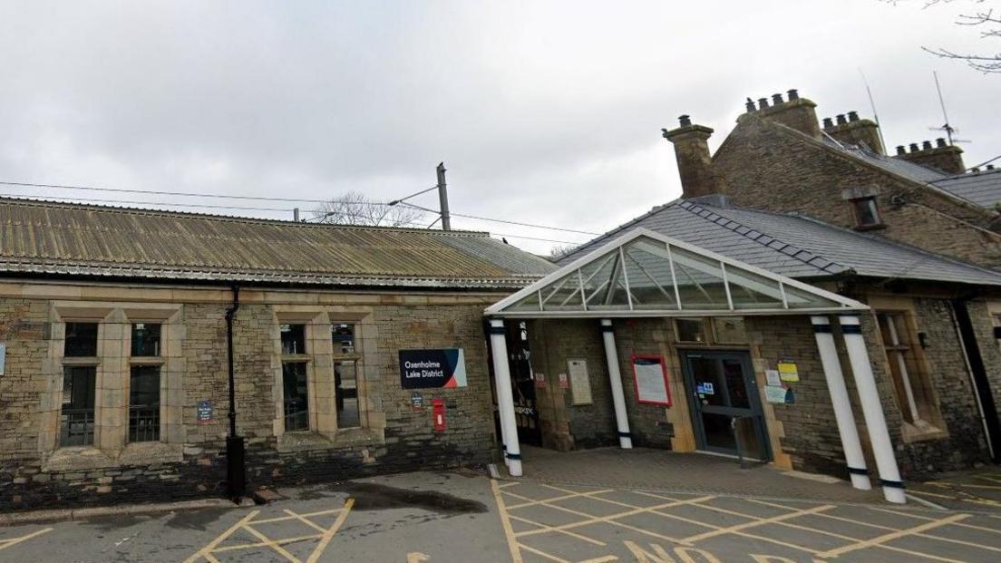 The entrance to Oxenholme train station. It is a one-storey stone building. There is a sign reading Oxenholme Lake District next to the entrance. 