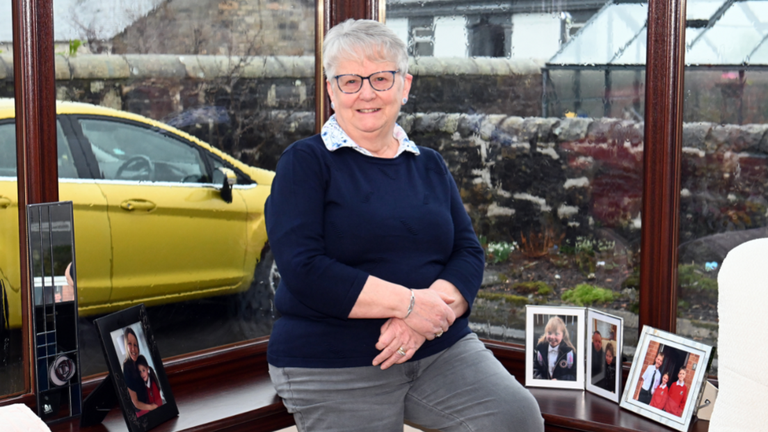 A woman perches on the windowsill of her bay window, wearing a navy jumper and grey trousers. There are framed photographs of her children and grandchildren around her and a yellow car is visible through the window.