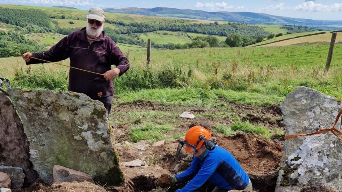 Two people work to measure and place the slate shards in the ground, which is dug up. The terrain is in a farming area in Jackson Ground, the Lake District.