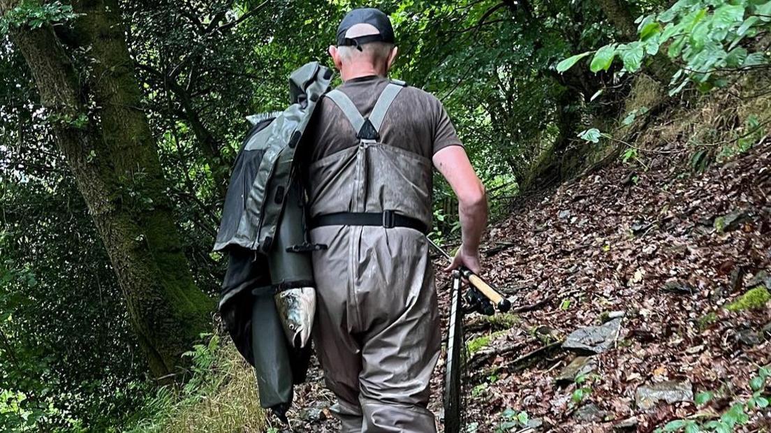 Stephen Samuel, wearing dungarees, is seen from behind with a whole salmon protruding from the sleeve of a coat slung over his shoulder