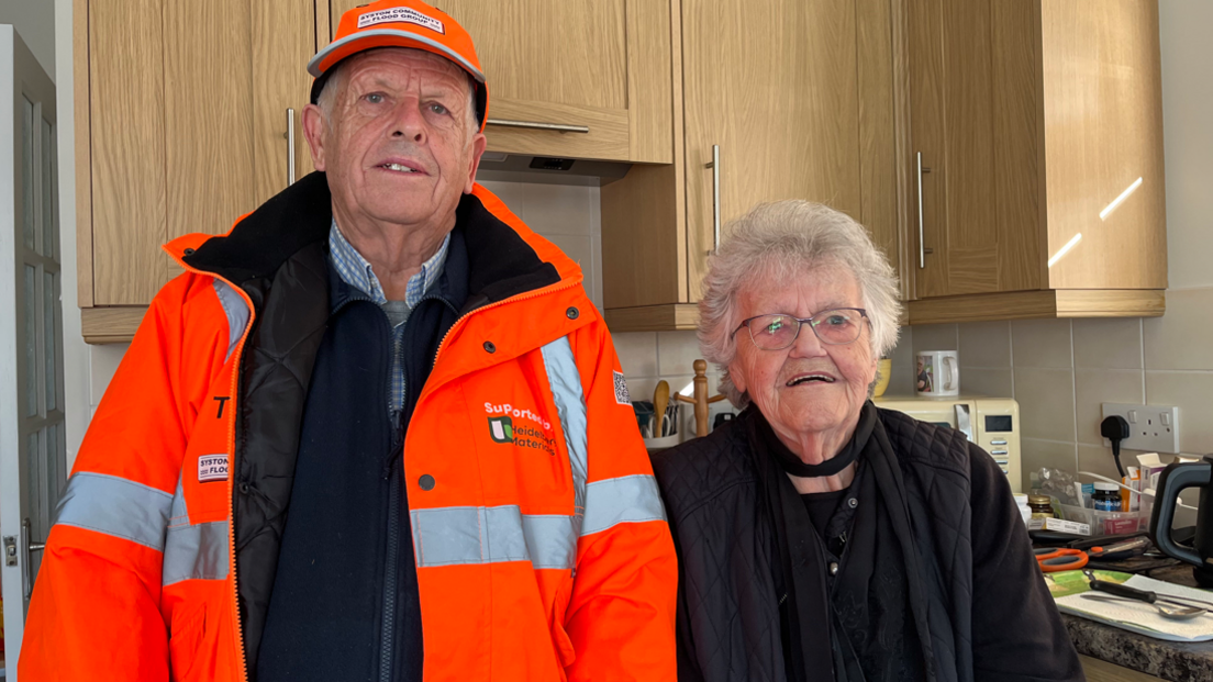 A man in a florescent flood warden jacket and cap with an older lady dressed in black