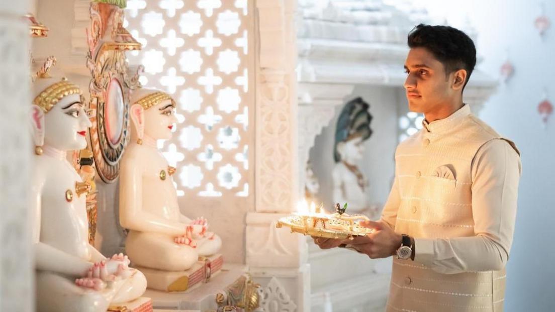 Dev standing on the right wearing a cream coloured kurta or traditional Indian top. He is praying while holding a plate with offerings and a diva in front of two white statues.