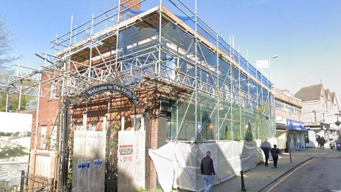 A brick building on a high street surrounded by scaffolding and cloth coverings.