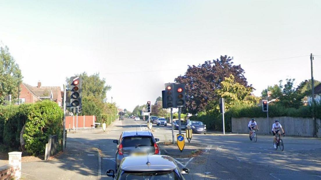 Two cars waiting at traffic lights at a road junction. Two cyclists are coming towards the camera. There are cars waiting to move in the opposite direction. There are trees and houses in the background.