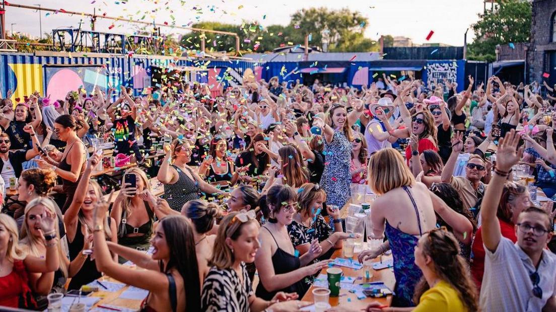 Dozens of people cheer as colourful confetti flies overhead during a Bingo Lingo session in the outdoor yard at Motion in Bristol