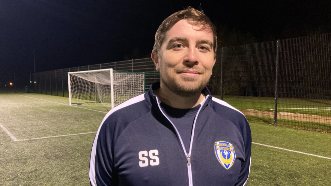 Sam Smith in navy club tracksuit with his initials SS on the front and an emblem of the football club. Sam is smiling at the camera and he has a short beard. There is a football pitch and a goal behind him.