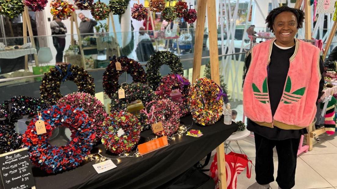 A woman dressed in black and a pink gilet with green Adidas logo standing next to a stall of floral and foliage Christmas garlands and wreaths. There are also multi-coloured floral pom poms hanging form the stall. Other traders can been seen by stalls in the building behind her.