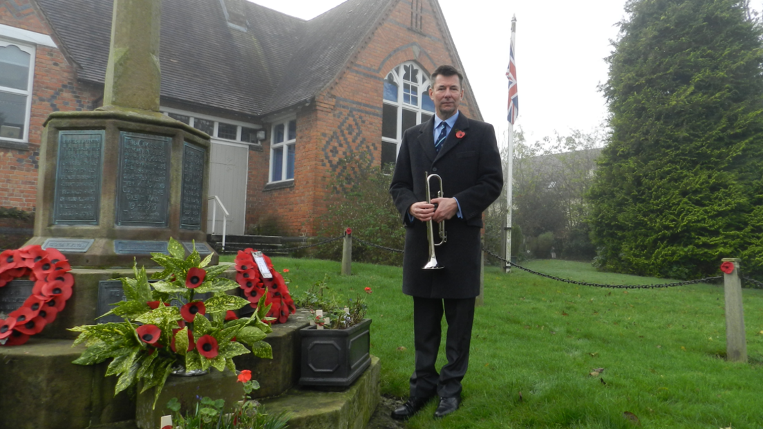 A man in a dark coat and suit and poppy, holding a bugle, standing beside a memorial with poppies on it. Behind him is a red brick building with some darker bricks in a diamond pattern. A big tree is to the right of the image with a flag pole with the union flag on it, next to it.