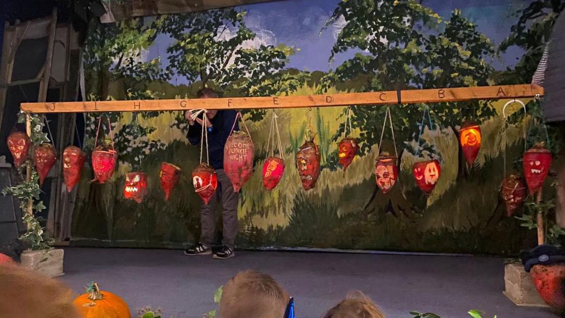 A row of Mangelwurzel lanterns is hanging off a wooden rail on a stage at Hinton St George watched by a group of school children.