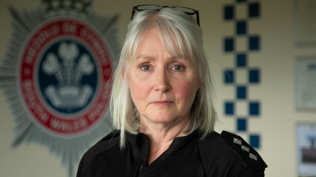 A middle-aged woman, Det Ch Insp Donna Clutterbuck, looks at the camera wearing police uniform standing in front of a South Wales Police logo on a wall