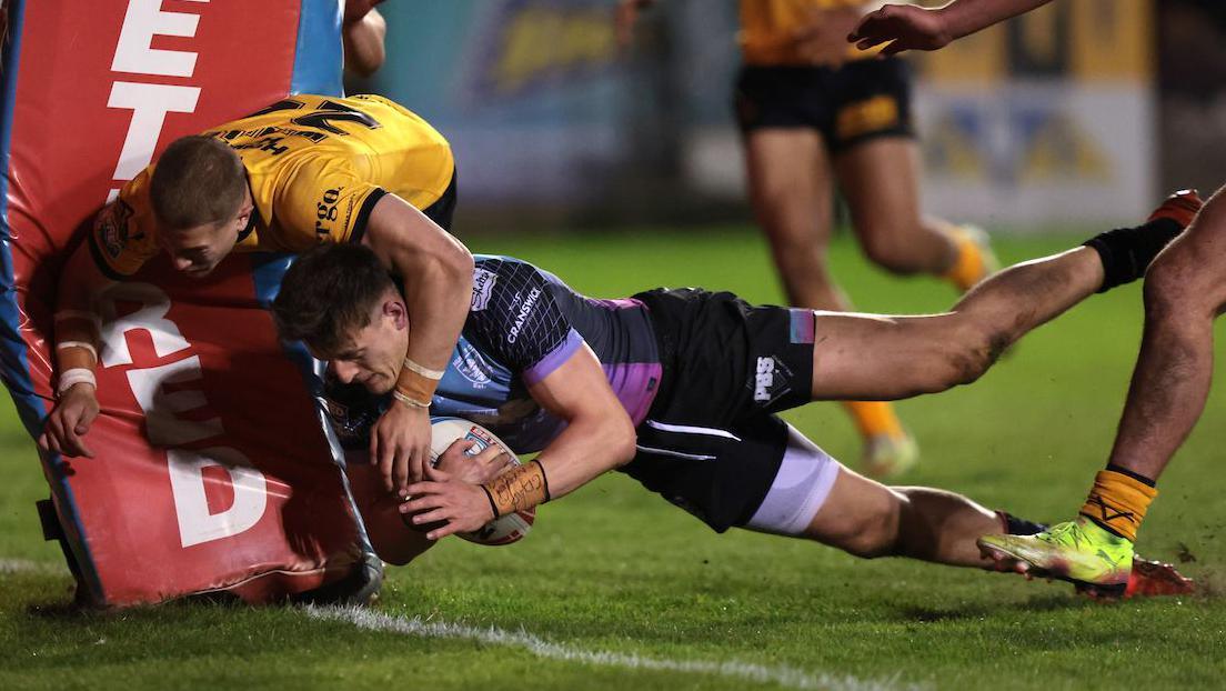 Hull FC's Davy Litten, in purple and blue shirt and black shorts, dives to score a try next to the post as Castleford Tigers' Jenson Windley, in black and amber, collides with the post while trying to stop him.