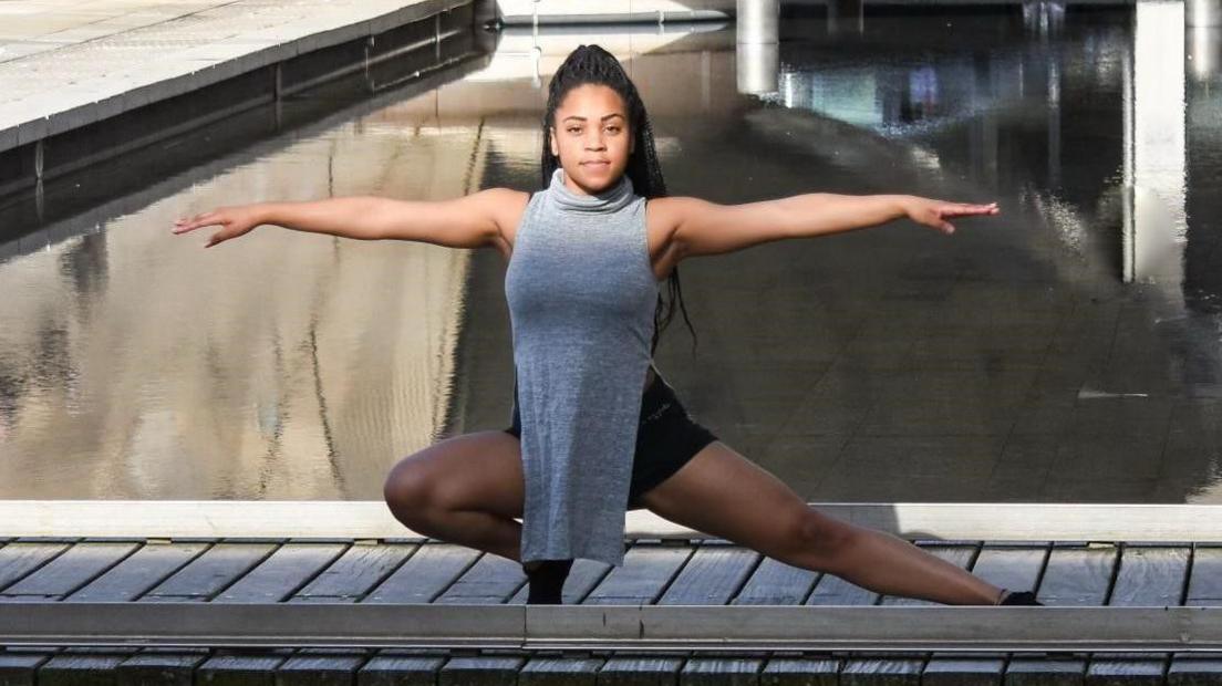 Candice Scott posing squatting near the floor with her arms out wide near some water