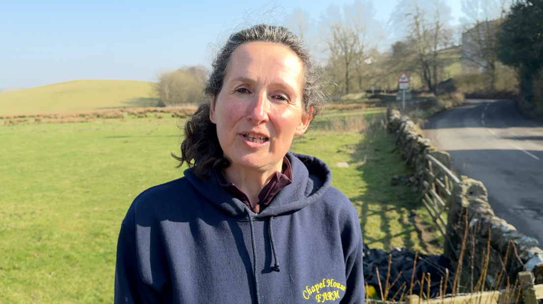 Jane McDaid standing in front of fields. She has dark hair tied back and wears a blue hoodie with a yellow logo saying Chapel House Farm.