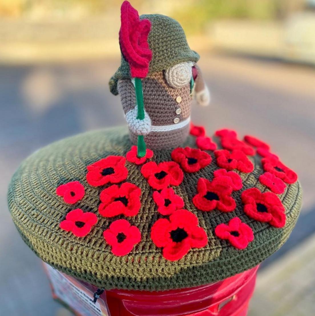 The knitted post box topper has red poppies and a figure dressed as a soldier who is holding a poppy.