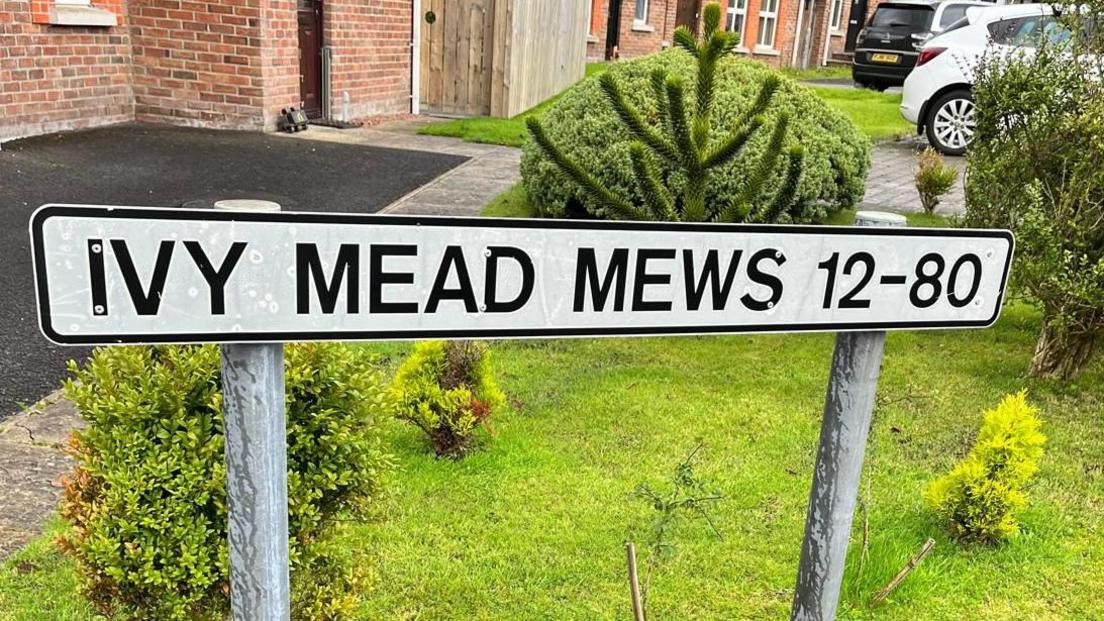 a street sign for numbers 12-80 Ivy Mead mews in Derry, standing outside a garden with a number of red brick houses in the background