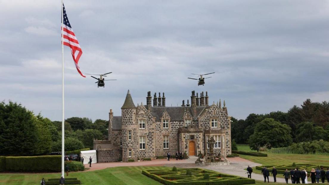 a small castle building beside a us a flag as two military helicopters fly overhead