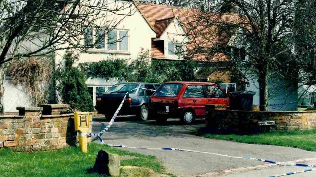 Police photograph of the front of the Brown's large farm house with a pitched chalet style roof. There are 2 cars in the drive and police tape cordoning the property off.