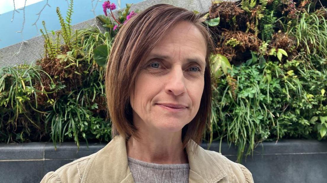 A woman with brown hair, a grey top and a beige jacket standing in front of a bush