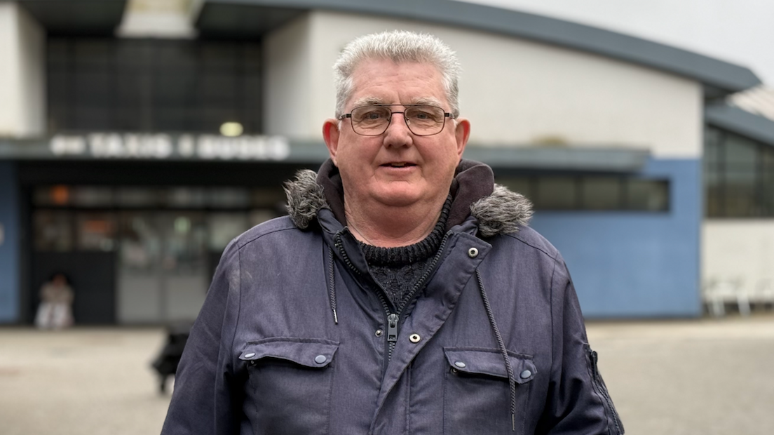 Billy Parker, has short white hair and wears black-rimmed glasses and a a purple and blue coloured jacket over a thick black jumper. A blue and white building is behind him, out of focus.